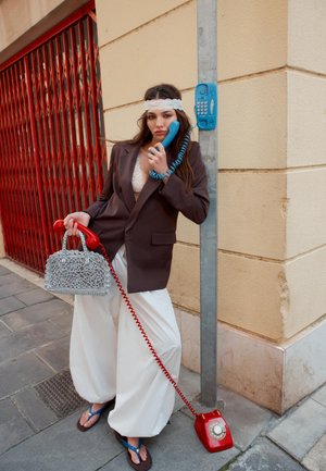 Kvinde i oversized blazer og hvide bukser holder en blå telefonrør fra en offentlig telefon til øret og en rød drejeskivetelefon med håndtaske i baggrunden.