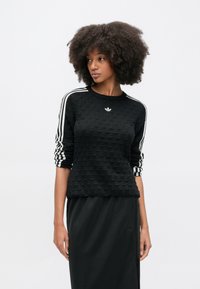 Čierna textúrovaná mikina s tromi bielymi vertikálnymi pásmi na rukávoch a malým vyšívaným logom. Kombinovaná s čiernou sukňou.