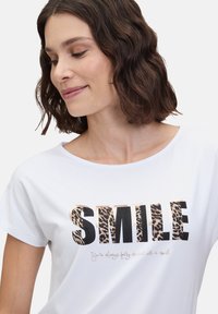 T-shirt blanc à manches courtes avec le mot "SMILE" en lettres noires grasses et imprimé léopard, accompagné d'un texte plus petit en cursive en dessous.