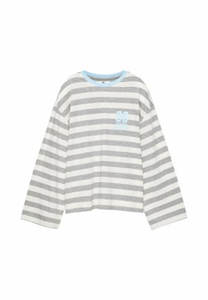 Jeune femme en t-shirt à manches longues rayé et jupe blanche assise sur une rambarde en pierre près d'une marina avec des bateaux sous un ciel bleu clair.