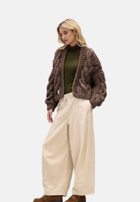 Brązowy dziergany kardigan z grubym wzorem warkocza, połączony z ciemnozielonym golfem i kremowymi szerokimi spodniami z sztruksu. Brązowe obuwie.