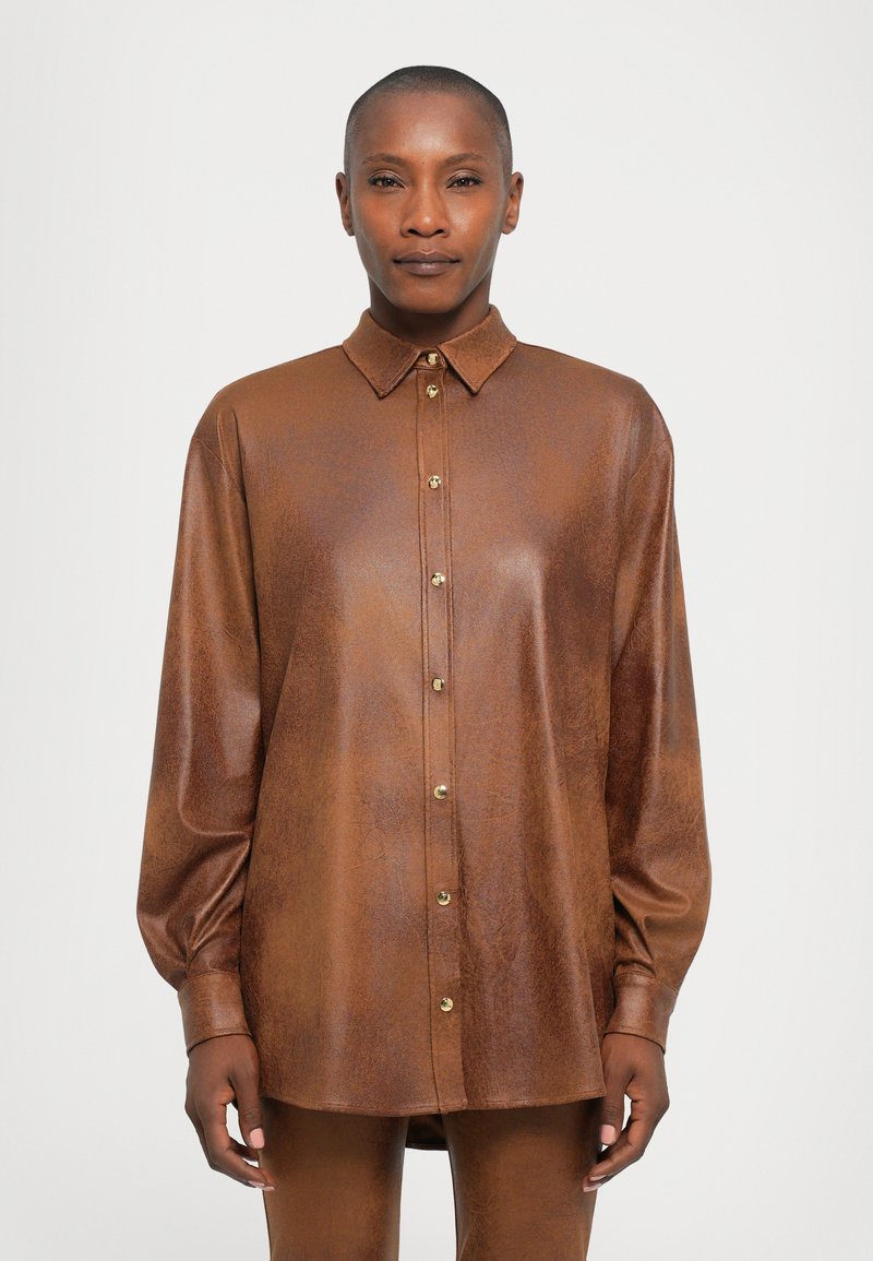 Person with short hair wearing a brown buttoned long-sleeve leather shirt and matching pants, standing against a plain background.