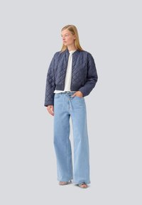 Veste bomber matelassée marine, coupe courte, poignets côtelés, portée sur un top blanc et un jean large bleu clair, avec une coupe décontractée.