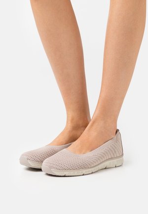 Pieds portant des chaussures décontractées beiges claires en maille respirante, sans lacets, avec semelles en caoutchouc flexibles sur fond blanc.