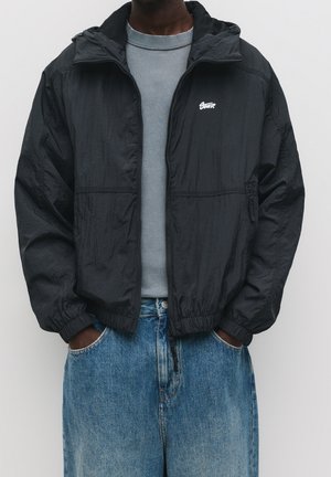 Haut du corps d'une personne portant une veste à fermeture éclair noire par-dessus un t-shirt gris, les mains dans les poches d'un jean bleu ample.