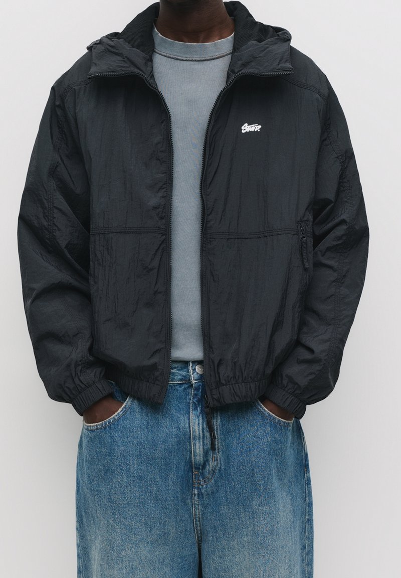 Haut du corps d'une personne portant une veste à fermeture éclair noire par-dessus un t-shirt gris, les mains dans les poches d'un jean bleu ample.