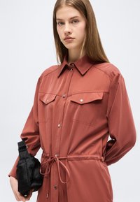 Νεαρή γυναίκα που φοράει ένα φόρεμα-πουκάμισο σε σκουριά απόχρωση, κουμπωμένο και εφαρμοστό στη μέση, κρατώντας μια μικρή μαύρη γούνινη τσάντα.