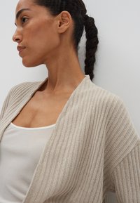 Lichtbeige, ribbelgebreide cardigan met een relaxte pasvorm, open voorkant en brede mouwen, gelaagd over een witte tanktop.