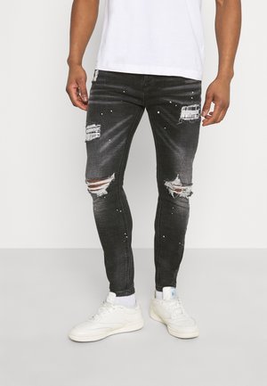 Hombre vistiendo jeans pitillo negros desgarrados con salpicaduras de pintura blanca, combinados con zapatillas blancas y calcetines sobre un fondo liso.