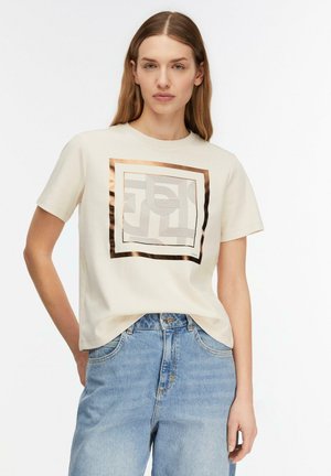 Donna con capelli lisci castano chiaro indossa una maglietta crema a maniche corte con motivo astratto di quadrati metallici e jeans a vita alta azzurri.