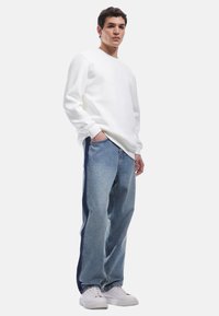 Sudadera blanca con cuello redondo, combinada con jeans azul claro que tienen paneles laterales más oscuros. La modelo lleva zapatillas blancas.
