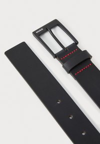 Ceinture en cuir noir avec une boucle en métal noire rectangulaire. Détails de surpiqûres rouges le long de la sangle. Présente plusieurs trous d'ajustement.