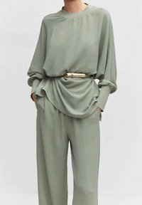 Blouse vert clair texturé à manches longues et coupe décontractée, assortie avec un pantalon identique, rehaussée par une ceinture dorée.