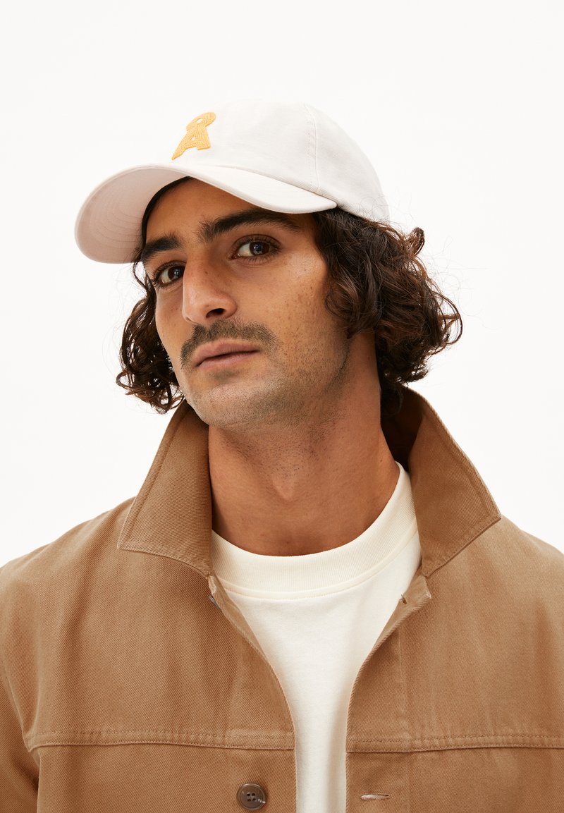 Witte pet met een gele geborduurde letter "A," gedragen met een bruine jas en een witte shirt. Krullend haar en een goed verzorgde snor zijn zichtbaar.