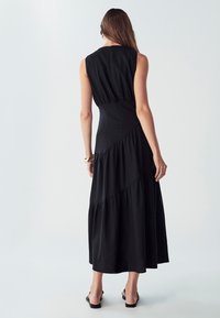 Črna brezrokavna maxi obleka s prilegajočim bodicem, večplastnim krilom in stranskim zadrgo. Tkanina ima gladko teksturo in subtilen sijaj.