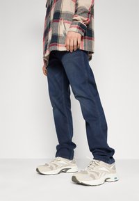 Jeans din denim albastru cu o tăietură dreaptă, asortați cu adidași texturați de culoare deschisă. O cămașă din flanel cu carouri în nuanțe de bej, roșu și bleumarin.