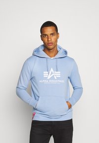 Felpa in pile azzurro chiaro con tasca a marsupio, logo bianco e cappuccio con cordino. Presenta polsini e orlo a costine, e una piccola etichetta rossa sul lato.