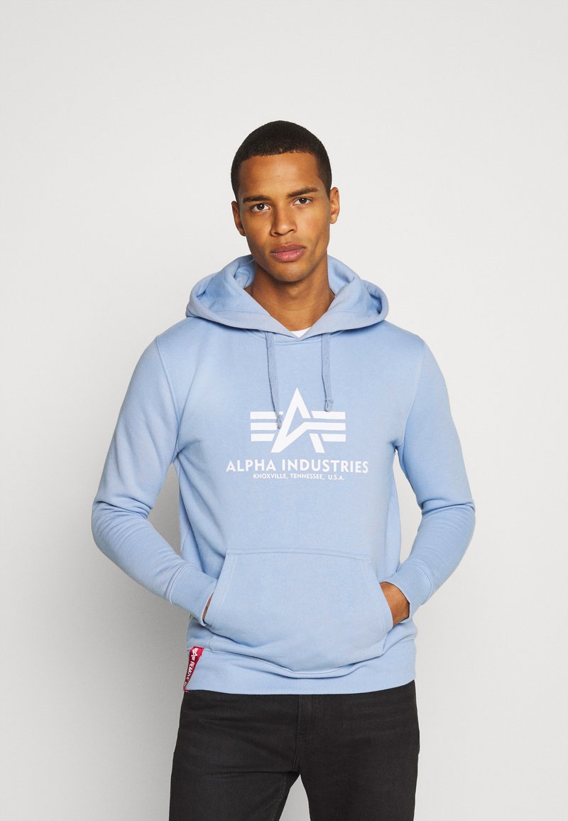 Felpa in pile azzurro chiaro con tasca a marsupio, logo bianco e cappuccio con cordino. Presenta polsini e orlo a costine, e una piccola etichetta rossa sul lato.