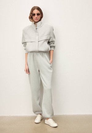 Szürke polár overall magas nyakkal és első zsebekkel, fehér tornacipővel párosítva. Simább textúra és kényelmes, bő szabás a kényelem érdekében.