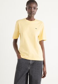 Gul bomulls-T-shirt med korta ärmar och rundhals, med en liten grön logotyp på bröstet. Kombinerad med mörka, löst sittande denimjeans.