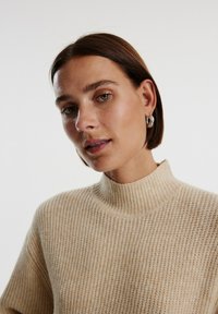 Beige gestrickter Pullover mit hohem Kragen, der über eine gerippte Textur und einen lässigen Schnitt verfügt. Das Model trägt silberne Ohrringe mit Blattdesign.