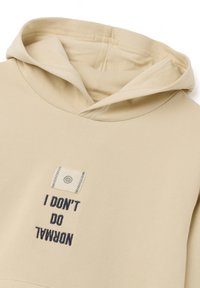 Hoodie beige avec une surface texturée, arborant une étiquette à l'avant qui indique "JE NE FAIS PAS DE NORMAL" en broderie bleu foncé.