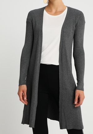 Mujer con un cárdigan largo gris acanalado sobre una camisa blanca y pantalones negros, de pie frente a un fondo claro y liso.