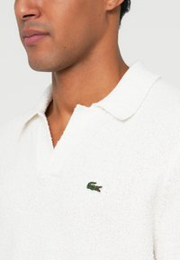 Chemise de col blanc en tissu texturé, avec un col court et un petit logo de crocodile vert sur la poitrine.