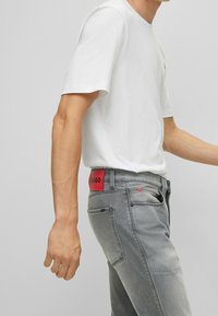 Vaqueros de mezclilla grises con una cinturilla de marca roja, bolsillos laterales y un diseño de pierna recta, combinados con una camiseta blanca ajustada.