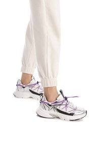 Zapatillas deportivas en blanco, negro y plata con cordones morados. Paneles texturizados, suelas gruesas y acentos de tacón prominentes. Diseño casual.