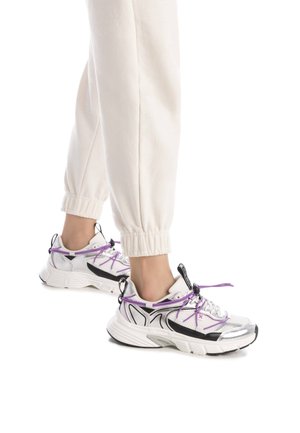 Zapatillas deportivas en blanco, negro y plata con cordones morados. Paneles texturizados, suelas gruesas y acentos de tacón prominentes. Diseño casual.