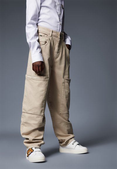 Pantaloni cargo beige con grandi tasche laterali, abbinati a sneakers bianche con un dettaglio di personaggio dei cartoni e logo sulla fibbia.