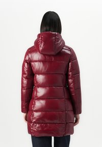 Vrouw in een glanzende, gewatteerde bordeauxrode pufferjas met een capuchon, staande met de rug naar de camera tegen een witte achtergrond.