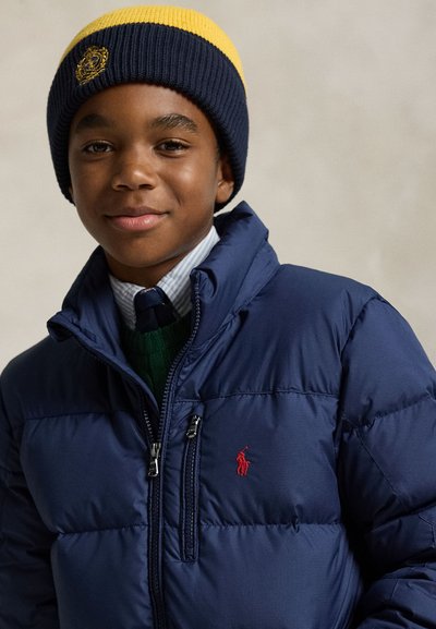 Băiat purtând o geacă matlasată bleumarin cu logo roșu, cămașă în dungi, cravată, pulover verde și o căciulă tricotată bleumarin și galben cu emblema brodată.