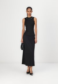 Top noir sans manches avec une finition texturée, associé à une longue jupe noire. Les accessoires comprennent un petit sac noir et des escarpins noirs à talons pointus.