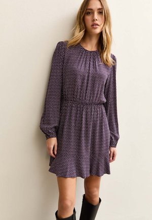 Vrouw draagt een lange mouwen, donkerpaarse jurk met polkadots, met een geplooide taille en zwarte kniehoge laarzen, staand tegen een effen muur.