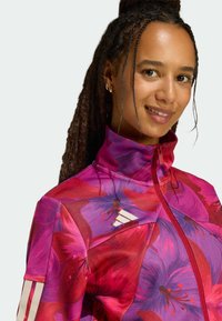 Veste à motif floral dans des teintes vives de rose et violet, avec un col montant, une fermeture éclair intégrale et des accents de rayures blanches sur les manches.