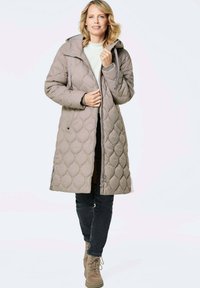 Hellbeige gepolsterte Jacke mit Kapuze und gestepptem Muster. Verfügt über einen Reißverschluss vorne und Kordelzüge. Getragen mit dunklen Jeans und hellbraunen Stiefeln.