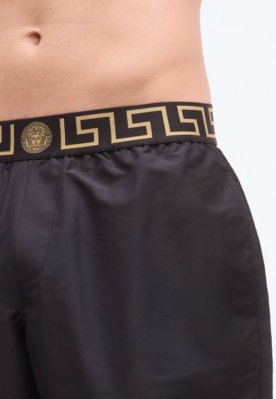 Zwarte zwembroek met een goud-zwart Grieks sleutelpatroon op de tailleband, met een Medusa-embleem centraal aan de voorkant.