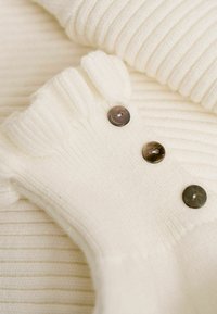 Witte ribbelgebreide kleding met drie donkere knopen, met een zachte textuur en een ronde halslijn. De stof lijkt gezellig en warm.