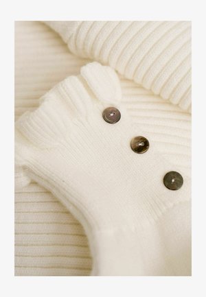 Weißes, geripptes Strickkleidungsstück mit drei dunklen Knöpfen, das eine weiche Textur und einen runden Ausschnitt aufweist. Der Stoff wirkt gemütlich und warm.