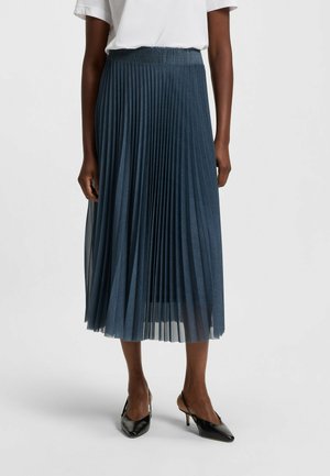 Frau trägt einen marineblauen plissierten Midirock, weißes kurzärmeliges Oberteil und schwarze spitze Slingback-Absatzschuhe, steht vor einem schlichten Hintergrund.