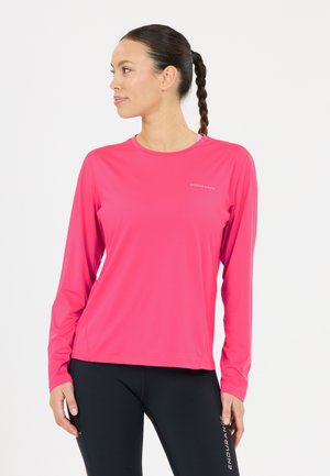 Frau mit dunklem geflochtenem Haar, die ein knallpinkes langärmeliges Shirt und schwarze Leggings trägt und nach links schaut vor einem schlichten weißen Hintergrund.