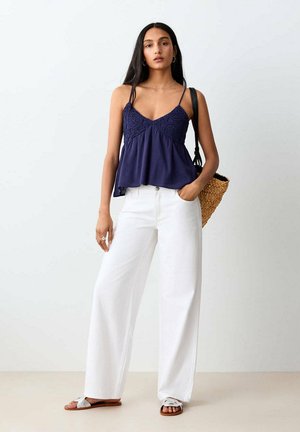 Femme portant un caraco en crochet bleu marine, un pantalon blanc à jambes larges, des sandales blanches, portant un sac en paille tressée sur l'épaule, debout devant un fond uni.