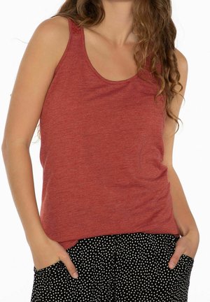 Femme portant un haut rouge sans manches et un pantalon noir à pois blancs, les mains placées de manière décontractée dans les poches du pantalon.