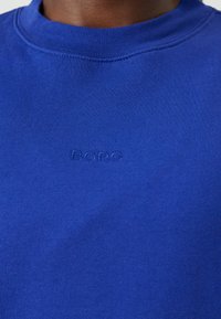 Felpa blu in cotone con scollatura tonda e logo ricamato "BOPC" dello stesso colore, esposta su un tessuto testurizzato.