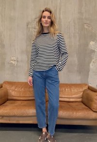 Naine seisab pruunist nahast diivani ees, kandes must-valget triibulist pika varrukaga särki, siniseid teksapükse ja pruune tossusid.