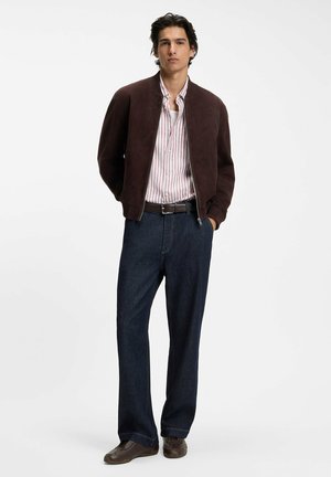 Mann steht mit den Händen in den Taschen, trägt eine dunkelbraune Bomberjacke, ein weiß-rot gestreiftes Hemd, dunkle Jeans und braune Schuhe.