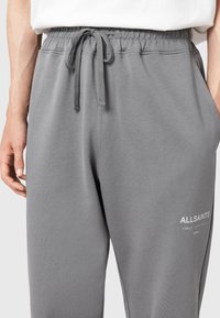 Pantaloni de trening gri cu șnur și talie elastică, cu logo-ul AllSaints pe coapsa superioară, purtați cu o cămașă albă băgată în pantaloni.