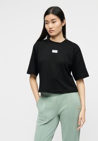 Νέα γυναίκα με μακριά μαύρα μαλλιά φοράει φαρδιά μαύρη μπλούζα και ανοιχτό πράσινο παντελόνι, στέκεται με το ένα χέρι στην τσέπη μπροστά σε λευκό φόντο.